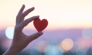 Sunset Colors Blurred Background to Person Holding Up Small Red Heart with One Hand