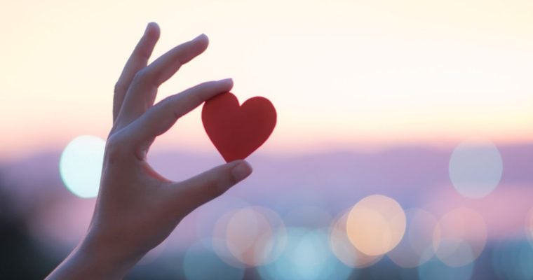 Sunset Colors Blurred Background to Person Holding Up Small Red Heart with One Hand