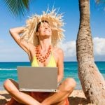 Smiling Woman Sits on Sand At Beach Under Palm Tree with Laptop