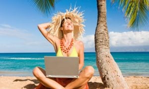 Smiling Woman Sits on Sand At Beach Under Palm Tree with Laptop