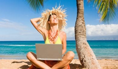Smiling Woman Sits on Sand At Beach Under Palm Tree with Laptop