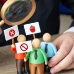 Man with Magnifying Glass Looks At 3 Small Human Figures Holding Protest Signs