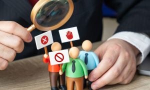 Man with Magnifying Glass Looks At 3 Small Human Figures Holding Protest Signs