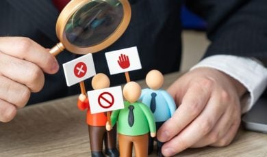 Man with Magnifying Glass Looks At 3 Small Human Figures Holding Protest Signs