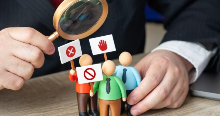 Man with Magnifying Glass Looks At 3 Small Human Figures Holding Protest Signs