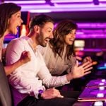 Bright Pink Lights of Slot Machines Shine While 3 Players Gambline Inside Casino