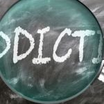 Magnifying Glass Sits on Blackboard with the Word Addiction Written