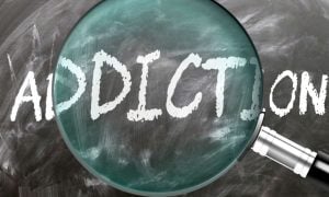 Magnifying Glass Sits on Blackboard with the Word Addiction Written