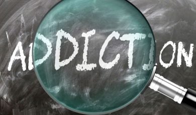 Magnifying Glass Sits on Blackboard with the Word Addiction Written