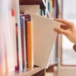 Woman Grabs White Book from Shelf Inside Bright Library