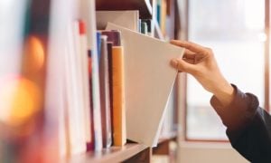 Woman Grabs White Book from Shelf Inside Bright Library