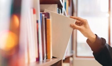 Woman Grabs White Book from Shelf Inside Bright Library