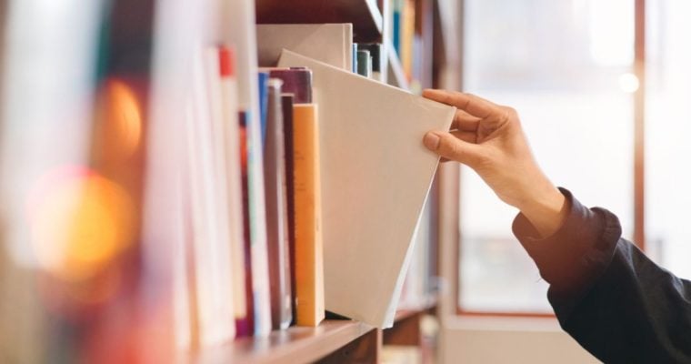 Woman Grabs White Book from Shelf Inside Bright Library