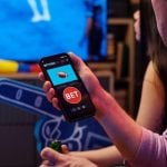 Two Young People Watch Football Game Inside, One Holds Phone with Sports Betting