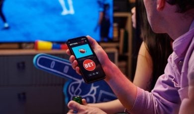 Two Young People Watch Football Game Inside, One Holds Phone with Sports Betting