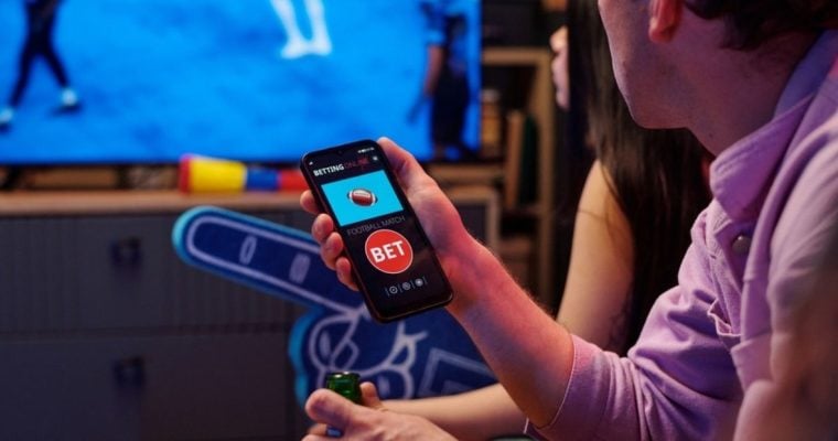 Two Young People Watch Football Game Inside, One Holds Phone with Sports Betting