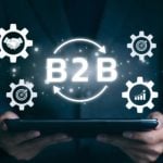 Digital Icons in White with Text B2B Hovers Over Tablet Held By Man in Dark Suit
