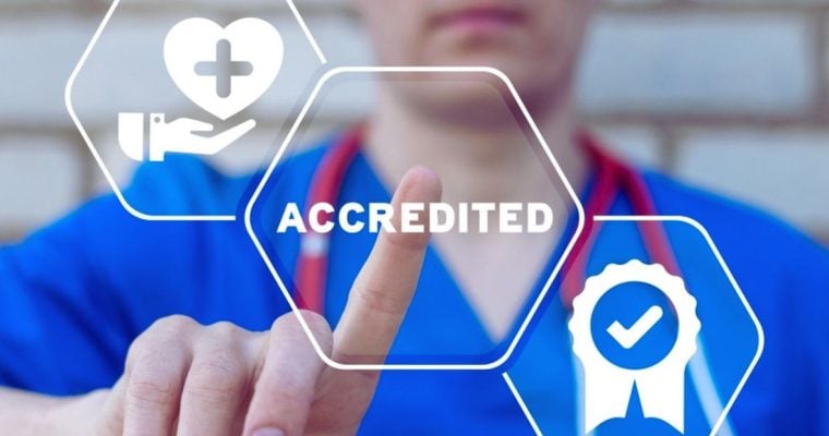 Hospital Nurse in Blue Garb Points to Digital Overlay of the Word Accredited in White Text
