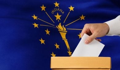Indiana State Flag Sits Behind Ballot Box with Man Placing White Envelope Inside