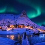 Man Stands On Snowy Bridge Looking at Village with Aurora Borealis Overhead