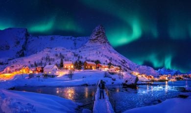 Man Stands On Snowy Bridge Looking at Village with Aurora Borealis Overhead