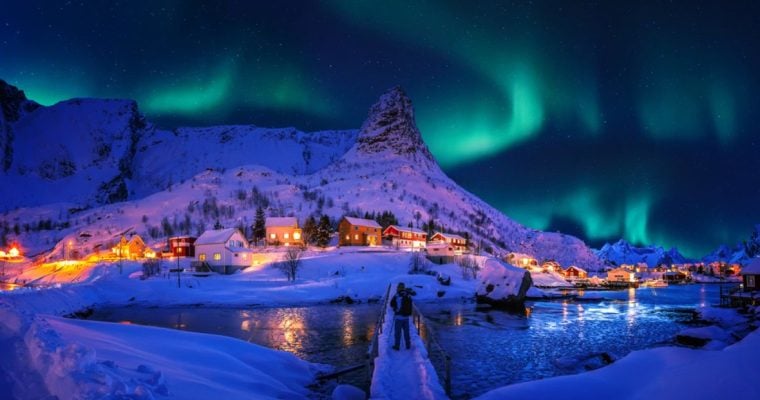 Man Stands On Snowy Bridge Looking at Village with Aurora Borealis Overhead
